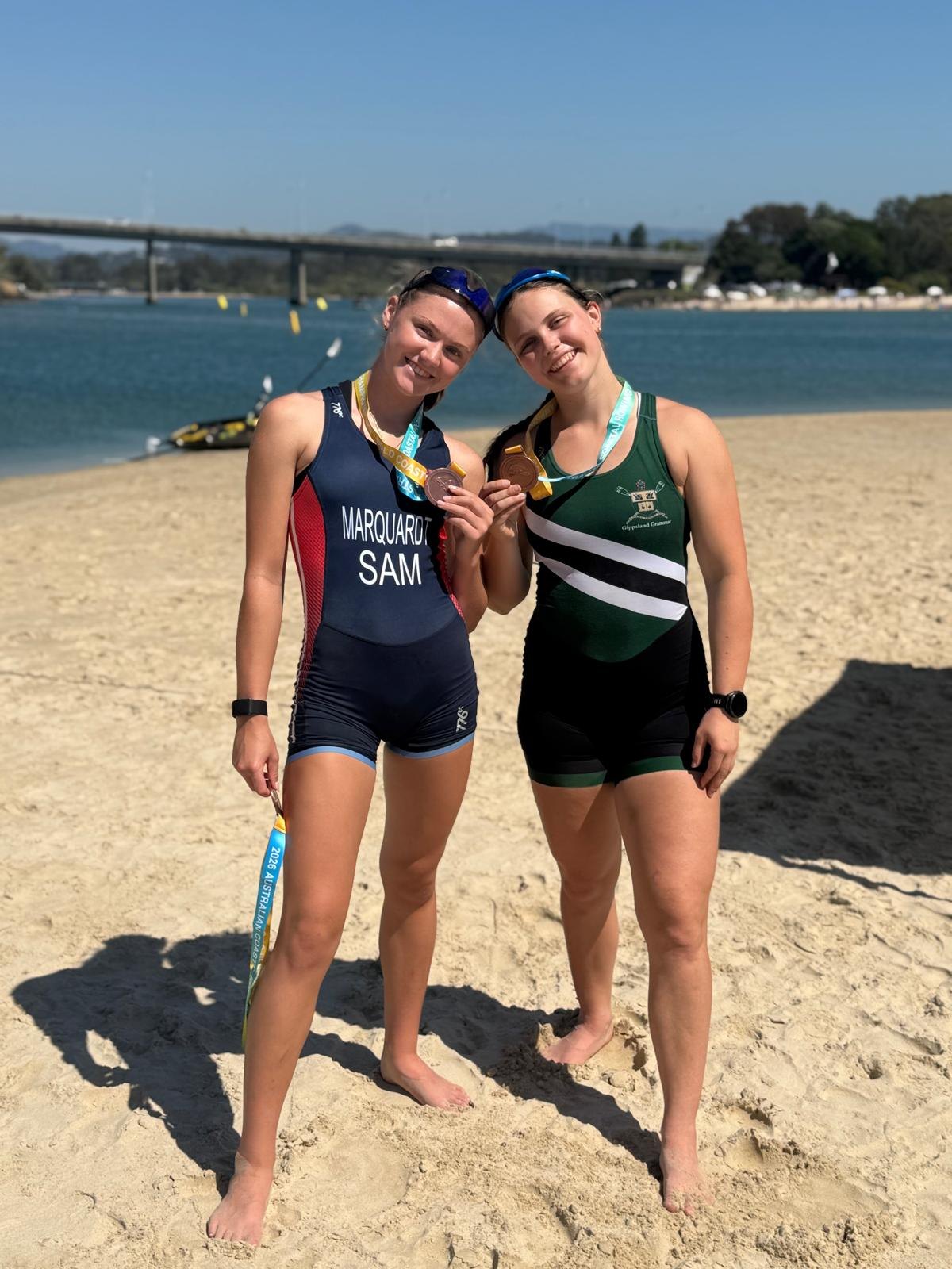 Alia Marquardt in the Samoa unisuit and Lily Canfield in Gippsland Grammar colours, standing on the sand at Currumbin Creek holding up their bronze medals