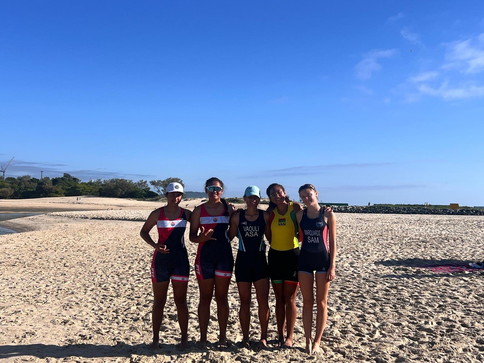 Five young women in the racing kit of French Polynesia, American Samoa, Vanuatu and Samoa, arms around each other on the sand at Currumbin Creek